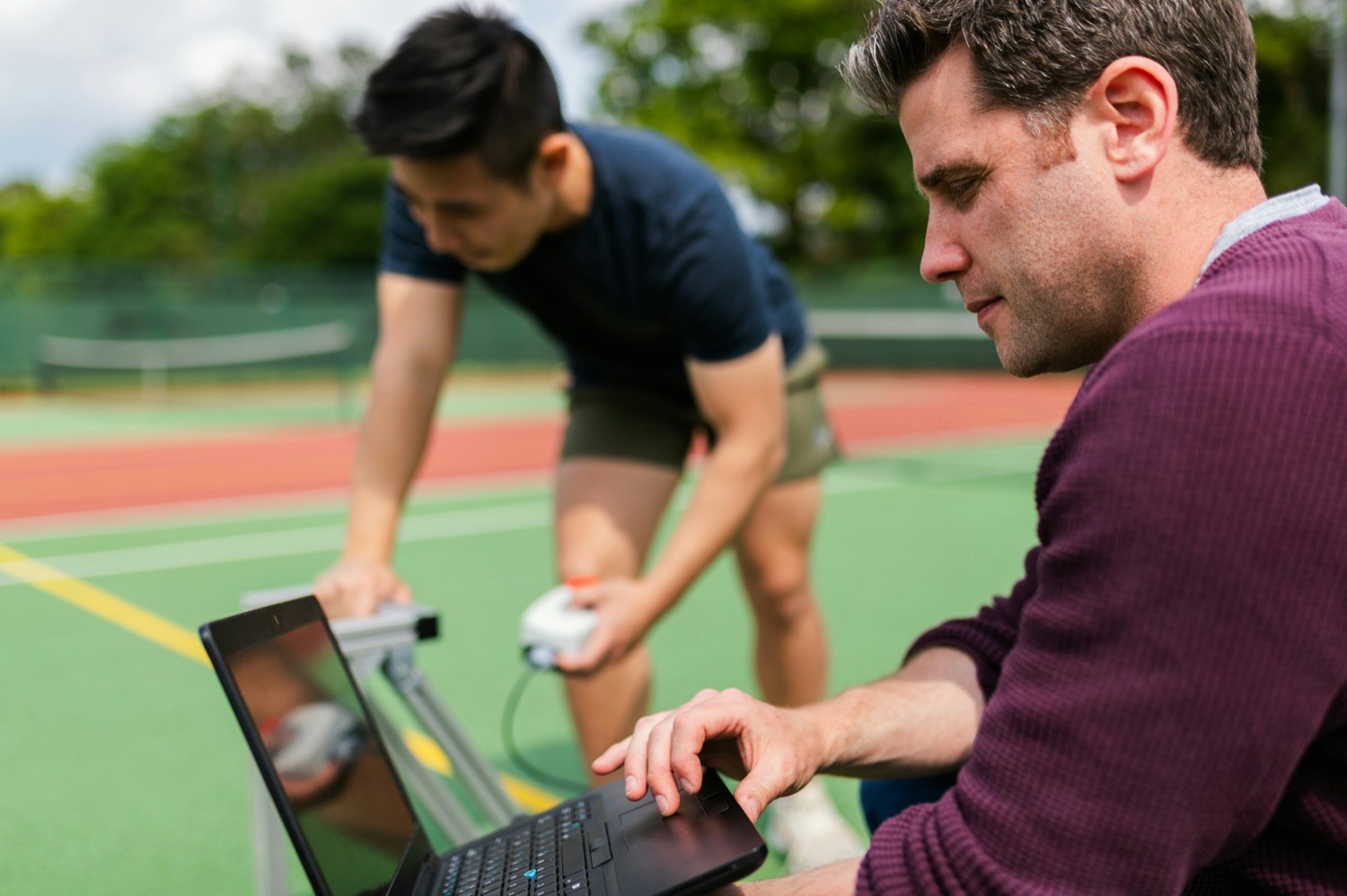 A clinician reviewing data on a laptop while an athlete is tested on a court behind him